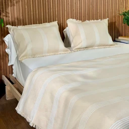 A single cream-colored cotton coverlet with a soft, striped pattern runs lengthwise across the bed, paired with matching pillow shams that feature delicate fringe edges. The overall look is light, neutral, and cozy, suggesting a unisex or general audience. Visible materials appear to be cotton in a smooth weave, with beige-cream tones and pale white stripes for a simple, natural style.