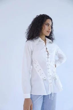 A white lace-up shirt with a relaxed, slightly oversized fit. It features long sleeves, a standard collar, and a crisscross lace-up detailing down the front and at the sides, giving a lace-up corset-inspired look. The material appears lightweight and semi-sheer, styled with light blue denim for a casual yet feminine vibe; intended audience: women.