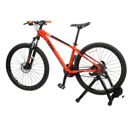 A red mountain bike is mounted on an indoor bicycle trainer stand, turning the bike into a stationary cycling setup. The visible model uses a full-suspension frame with thick knobby tires, orange handlebar grips, and a compact trainer frame supporting the rear wheel; pedals and chain are visible, indicating a classic 2x gear drivetrain. The setup appears suitable for adults (unisex), with emphasis on sturdy frame, black trainer legs, and front wheel stabilizer for indoor use.