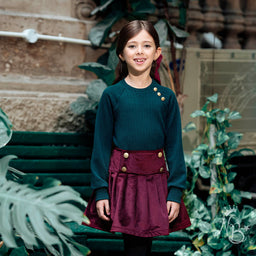 Description:
- A front-button burgundy skirt paired with a dark teal knit top. The girl is wearing a high-waisted, A-line skirt with prominent gold buttons along the front and sides, and a matching fabric belt detail at the waist visible from the side. The look is styled for a young girl.
Audience and style notes:
- Intended audience: girls (youth).
- Visible attributes: burgundy velvet-like fabric, gold-tone buttons, double-belted side detail, long-sleeve teal sweater, knit texture, and a knee-length, slightly flared silhouette.