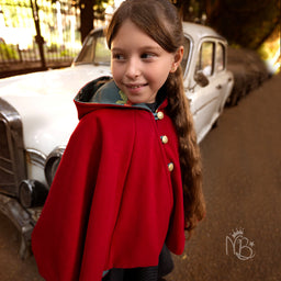A Christmas cape for a child, shown worn by a girl with a long braid. It appears unisex in cut but styled for a girl, with a bright red outer fabric and a dark lining visible at the hood, plus gold-tone buttons along the front. Notable attributes include a soft, wool-like red material, cape silhouette, hooded design, and a classic festive holiday look.