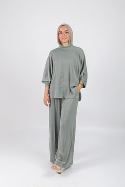 The photo features a loose, olive-green matching set consisting of a high-neck, oversized top with wide, three-quarter sleeves and wide-leg pants. The outfit appears designed for women, styled modestly with a light hijab in a complementary tone, and worn with neutral sandals. Notable attributes include the soft, flowy fabric, solid olive color, and relaxed, contemporary silhouette.