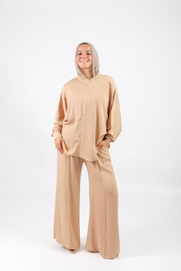 - The image shows a beige loungewear set consisting of an oversized hoodie and wide-leg pants. The model is wearing a relaxed, hooded top with large front pockets and matching flowy trousers, both in a soft, neutral tan shade.
- Audience: women (slightly oversized, drapey style and fit suggests female demographic).
- Notable materials and styling: soft, knit or fleece-like fabric with a smooth matte finish; monochrome beige color; loose, comfortable silhouette with a roomy hood and wide legs.