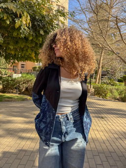 - The image shows a woman modeling a jacket worn open over a white cropped or fitted top and high-waisted jeans. The jacket appears color-blocked, with solid black on the upper portion and blue patterned denim or velvety blue panels on the lower sleeves and sides.
- Intended audience: women's apparel inferred from the styling, fit, and proportions.
- Notable attributes: black upper body, blue patterned lower panels, zip or button front (hidden by open wear), casual/athleisure streetwear vibe, mid-weight jacket suitable for cool weather. Material hints suggest a mix of black fabric with blue denim-like or textured panels, and a relaxed, slightly oversized cut.
