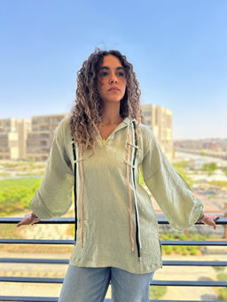 - What the product is: A light-green, long-sleeve top with a loose, relaxed silhouette and a textured fabric. It features a polo-style collar and a lace-up detail along the front.
- Apparent audience: Women (based on the fit, styling, and model).
- Notable attributes: Light sage/green color, textured soft fabric, voluminous balloon sleeves, extended hem, and decorative vertical ribbon/lace-up accents with contrasting darker tones. The overall look is casual, breezy, and flowy.