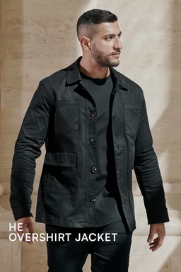 A men's shirt jacket (shacket) in a dark, nearly black color. The model wears a lightweight, structured jacket with a button-front closure, long sleeves, and a stand-up collar. Visible features include front pockets and a clean, minimal design with a casual, layered look suitable for outerwear in cooler weather.