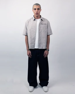 This is a short-sleeve, boxy-fit striped shirt in a beige and light neutral tone, layered over a white inner tee. It appears intended for a masculine or unisex audience, given the straight, loose cut and broad-shouldered silhouette. Visible attributes include a relaxed short sleeve, a collared neckline, vertical subtle stripes, and a small embroidered or stitched logo on the chest, paired with dark wide-leg pants and white sneakers for a casual, streetwear-inspired look.
