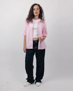 - What it is: A short sleeve linen shirt worn open over a white crop top, styled in a relaxed, boxy fit.
- Intended audience: Women's (as indicated by the model and styling).
- Notable attributes: Dusty rose pink linen fabric, short sleeves, oversized/boxy silhouette, button-front shirt, paired with dark wide-leg pants and white sneakers. Visible styling shows a casual, breathable summer look.