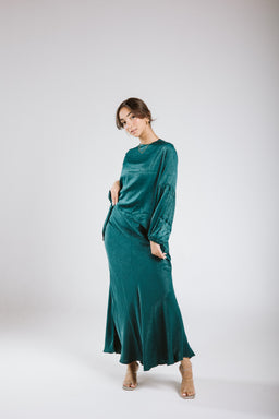 This is a teal, shimmery satin two-piece set (top and long skirt) worn by a female model. The audience appears to be women; the styling is elegant and flowy with a relaxed-fit long-sleeve top and a high-waisted, ankle-length skirt. Notable materials and attributes visible: satin/shiny velvet-like fabric in teal, with a soft drape and a subtle sheen; both pieces have a coordinated, cohesive look suitable for dressy or evening wear.