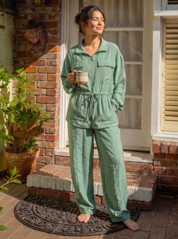 - A woman stands on a porch wearing a mint green linen matching set (top with a collar and long sleeves, drawstring waist pants). She is barefoot and holding a mug, giving a casual, relaxed vibe.
- Audience: appears unisex/ladies’ loungewear; styling suggests women’s sleepwear or lounge set. Notable attributes: light mint green linen, relaxed fit, collared button-up top with pockets, elastic/drawstring waist on wide-leg pants.