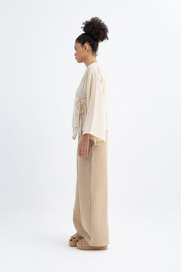 Description: A linen lounge set in light cream and dark beige tones worn by a female model. The ensemble includes a loose, draped top with wide sleeves and a waist-tie, paired with wide-leg pants. The look is relaxed, neutral, and breathable, with a natural, textured linen fabric. Audience: women's. Notable attributes: light cream top, dark beige/khaki pants, matching belt/tie at the waist, oversized silhouette, open-toe sandals. Material: linen; Style: casual, loungewear or resort set.
