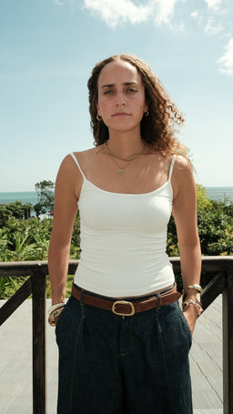 A white spaghetti strap tank top on a female model, styled casually outdoors with a wooden railing and greenery in the background. The top is fitted, sleeveless with thin shoulder straps, and appears made of a lightweight knit or jersey. The look is simple and summery, paired with dark high-waisted jeans and a brown belt for a clean, everyday outfit.