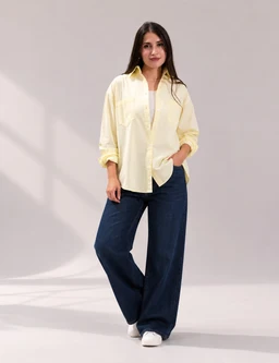 A yellow poplin shirt, styled loosely with a relaxed fit and rolled sleeves. It’s worn by a female-presenting model, suggesting a unisex-to-women audience, with a casual, oversized silhouette. Notable features include the soft pastel yellow color, button-front placket, and a crisp poplin fabric paired with wide-leg dark jeans.