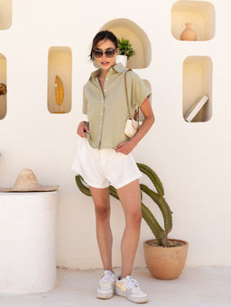A linen shirt worn by a female model. Appears intended for women (the styling is feminine with a relaxed, casual vibe). The shirt is light sage/khaki in color with short sleeves, a collar, and button front; paired with white shorts, white sneakers, and a small crossbody bag for a breezy, warm-weather look. Visible materials emphasize lightweight linen/natural fiber, soft drape, and a casual Summer vibe.