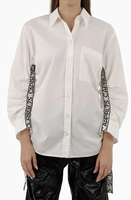 - What it is: A white button-down shirt with a loose, relaxed fit, and long sleeves. It features graphic tape-like detailing along the outer sleeves with text repeated along the seam.
- Intended audience: Unisex-or-women depending on fit; the styling with a slightly oversized silhouette and the model suggests a women’s fashion look.
- Notable attributes: Crisp white cotton/soft fabric, classic shirt collar, front button closure, and bold black tape accents running along the outer sleeves with repeated text. Overall style is contemporary, sporty-chic with a slightly oversized, casual vibe.