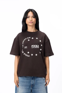 - The image shows a brown oversized short-sleeve T-shirt with a graphic centered on the chest. It features white circular text and star motifs surrounding a logo area.
- Audience: unisex or general adults, suitable for both men and women, based on the relaxed fit and styling.
- Notable attributes: dark brown fabric, short sleeves, crew neck, graphic print in white with circular text and stars, casual streetwear style. Material appears to be cotton or cotton-blend jersey.