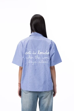 - Product: A light blue, short-sleeve button-up shirt with a relaxed fit. The back features white embroidered text reading “art is loude when the word stays silent” (likely stylized).
- Audience and styling cues: Designed for a unisex or women’s/teen audience based on the oversized cut and soft fabric; worn casually with jeans as shown.
- Materials and colors: Light periwal-blue cotton or cotton-blend fabric with a crisp texture; white embroidery provides contrast. Notable style: short-sleeve camp collar shirt with back embroidered message.