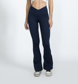 A pair of navy blue criss-cross yoga pants with a high, V-shaped waistband. The model appears to be a woman, shown from mid-torso to feet, wearing a white crop top; the pants hug the hips and thighs and flare slightly at the calves. Notable details include the dark blue color, smooth stretch fabric, and a snug, form-fitting fit with a subtle flare at the bottom hem.