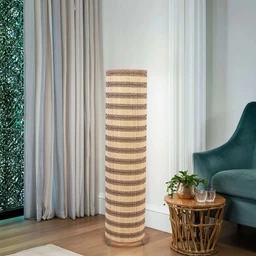 - A tall floor lamp with a woven rattan or wicker shade and stand, standing indoors on a wooden floor.
- Appears for a unisex or home décor audience; the design is natural and earthy, with alternating light and dark horizontal stripes running around the cylindrical shade.
- Notable attributes visible: cylindrical lamp shaft made of woven rattan, warm beige and brown tones, soft ambient light. The surrounding décor includes a small side table with a plant and glasses, a teal armchair, and light-colored curtains in the background.
