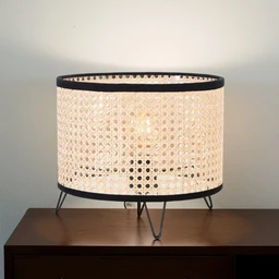 - A woven rattan lampshade with a drum shape, placed on a wooden cabinet. The shade has a light beige color with a perforated, circular pattern and dark trim along the top and bottom edges, supported by small metal legs.
- Intended audience: unisex (suitable for home decor for both men and women), judged from its neutral, minimalist styling and compact table-lamp form.
- Notable attributes: natural rattan material, light beige color, circular perforated weave, drum shade, black trim, tripod-like metal legs, compact 25×15 cm dimensions suggesting a small bedside or accent lamp.