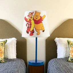 - Description: A table lamp with a white drum lampshade featuring cartoon characters (Winnie the Pooh and Tigger). The base and stem are bright blue, mounted on a round blue pedestal stand. The lamp is placed between two single beds with patterned bedding in a bedroom setting.
- Audience and attributes: Appears to be for a kids’ or youth bedroom (unisex), suitable for children due to playful characters and colorful design. Notable materials and style: fabric lampshade, solid-colored ceramic/metal base in blue, simple modern silhouette with a whimsical print. Color palette includes white shade, blue base, and multicolor characters. Size implied by title: approximately 23x45 cm.