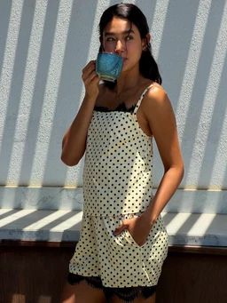 - What the product is: A light, sleeveless romper dress in a polka dot pattern, with a layered short length and a relaxed fit; the model holds a blue mug in the photo.
- Apparent intended audience: Women (adult female model, styling and fit suggest women's loungewear or casual sleepwear).
- Notable attributes: Cream/ivory base with black polka dots, black trim along the neckline and hem, side pockets, lightweight fabric, and a casual camisole-style top with thin straps.