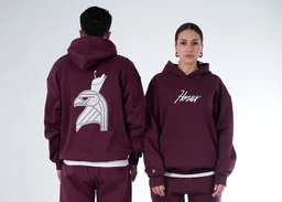 A matching hoodie set in a deep cherry/maroon color. The model (unisex styling) wears a pullover hoodie with a front script logo on the left chest and a large graphic of a helmeted figure on the back; the back view also shows an oversized print. Notable materials appear to be soft fleece/sweatshirt fabric with ribbed cuffs and hem, and a white inner layer peeking at the waist, suggesting a layered, casual streetwear look.