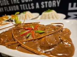 - What the product is: A plate of steak in a rich brown pepper sauce, with decorative fried onion strings and a small herb garnish.
- Apparent audience: Unisex (suitable for both men and women) judged from the plating and rustic steak presentation.
- Notable attributes: Visible glossy brown pepper sauce over sliced steak, thin fried onion strings arranged on top, small red and green pepper pieces and herbs as garnish, and a white plate with a blurred background. No visible branding.