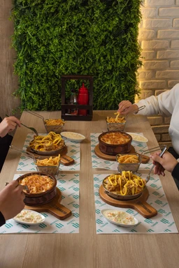 - What the product is: A rustic-style set of hot dipping dishes featuring bread bowls filled with warm cheesy dip and a side of fries; multiple servings arranged on wooden boards with small bowls of sauces.
- Apparent audience and styling: Appears to target adults dining in a casual, shared-meal setting; unisex presentation with hands of different people visible, suggesting group or family use. The scene suggests a party or casual gathering vibe.
- Notable attributes: Visible ingredients include French fries and melted cheese or dip inside small round bowls on wooden boards. Colour palette features warm browns from the boards and bowls, golden fries, and a cheesy yellow-orange dip; background includes a green plant wall and light brick, contributing to a cozy, rustic dining aesthetic.