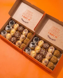- What the product is: A sharing box containing an assortment of small donuts arranged in two open orange boxes.
- Apparent audience: Unisex, suitable for adults and older teens given the neutral packaging and common donut variety.
- Notable attributes: Visible donut varieties with different coatings (chocolate, powdered sugar, and nutty/crunch coatings) in a light brown color palette; two identical boxes with a simple, warm-toned orange backdrop; no branding visible beyond generic text on the inner box lids.