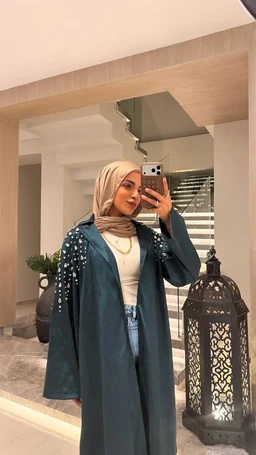 - The product appears to be a long, teal-green outer coat or duster with decorative embellishments along the sleeves or shoulders. It is styled over a light top and jeans, paired with a beige hijab, photographed as a street-wear or fashion look.
- Audience: women (universal feminine styling indicated by hijab, coat, and overall cut).
- Notable visible attributes: teal/greenish coat with pearl or bead embellishments on the upper sleeves/shoulders, long loose silhouette, wide sleeves, layered over a light top and high-waisted jeans. The setting is a fashion look with a modern, modest style.