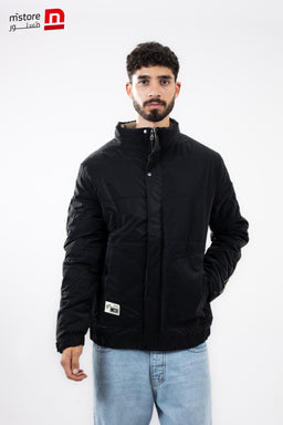 The product is a black waterproof jacket with a high collar and zip closure, designed for cold or wet weather. The model appears to be a man, suggesting a men’s fit, with a casual, urban styling. Notable details include a quilted padded construction, a front zipper concealed by a placket, side pockets, and a small label patch near the hem. The jacket has a matte finish and a minimalist, versatile look suitable for everyday wear.