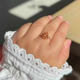 - A delicate ring worn on the left hand’s ring finger, featuring a peachy-orange central gemstone with smaller clear stones on its band. The model’s pink-tinted nails and the white lace sleeve frame the ring. 
- Intended audience appears to adults (unisex look but styled for women), judging from the delicate gemstone, thin band, and feminine setting. 
- Notable attributes: warm peach/orange central gemstone, small clear side stones, slender metal band, vintage-inspired prong setting, overall soft, romantic style.