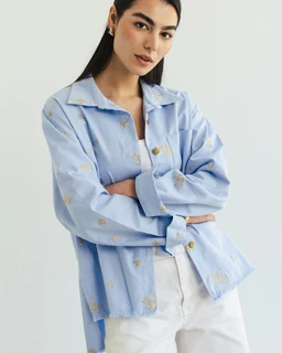 - A light blue, long-sleeve button-front shirt/jacket with a relaxed fit, worn open over a white top and white pants. The garment appears to have subtle embroidery or faded floral/pineapple-like motifs in a pale neutral color.
- Target audience: unisex or women’s, judged from the model styling and slightly oversized cut.
- Notable attributes: sky blue denim-like fabric, pastel embroidery, classic shirt collar, chest pocket, and silver-tone buttons; overall casual, airy, and soft-toned with a minimal, summery vibe.