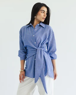 The garment is a long-sleeve button-up shirt in blue with white vertical stripes, shown tied at the waist to create a wrap/knot detail. Appears targeted toward adult women, with a relaxed, slightly oversized fit and rolled sleeves. Notable materials visible suggest lightweight woven cotton or a cotton-blend; colors are sky blue and white stripes with a soft matte finish, classic collar, and a waist-tie belt for a wrapped silhouette.