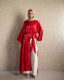 - Product: A red, long-sleeve open-front robe or abaya-style outerwear with a matching waist tie. The garment is worn over white wide-leg pants and paired with a light-colored headscarf.
- Audience and styling cues: Designed for women, suggested by the hijab styling, modest cut, and robe-like silhouette. The look is elegant and modest, suitable for everyday wear or semi-formal occasions.
- Materials and colors: Bold cherry-red fabric with a satin-like sheen; white wide-leg pants visible beneath; neutral beige or taupe headscarf. The belt ties at the waist create a gathered, draped effect. The overall style is flowy and contemporary modest fashion.