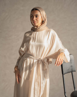 A modest, cream-toned abaya or long robe with a relaxed, flowing silhouette. It features a self-tabric tie belt at the waist and wide, draped sleeves, complemented by a matching hijab for a coordinated modest look. The fabric appears light and soft with a subtle sheen, giving an elegant, understated style.