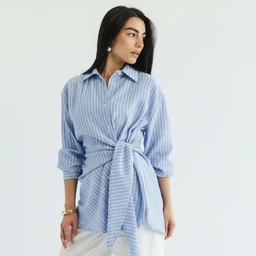 This is a light blue striped wrap shirt styled as a relaxed button-down, with a fabric belt tied at the waist for a cinched silhouette. The model appears to be a woman, suggesting a women’s or unisex fit with a casual, flowy cut. Notable details include the vertical stripes, long sleeves rolled up to the forearm, and a soft, breathable shirting material.