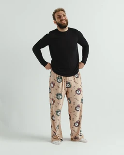 This is a pair of lounge pants (Alarms Fleece Pants) worn by a male-presenting model. The apparent audience is men’s, judged from the loose, relaxed fit and styling. Notable attributes visible: soft, fleece-like fabric in a light beige/peach tone with a repeated circular alarm-clock motif in darker colors; the pants have a relaxed, straight-leg cut and an elastic/comfortable waistband. The model pairs the pants with a black long-sleeve top, and the overall look conveys a cozy, home-loungewear style.