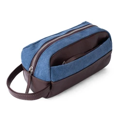 A compact Dopp kit-style bag with a two-tone blue denim-like upper and dark brown leatherette base and trim. It features a top zipper closure and a front exterior pocket, with a brown leather handle for handheld use. The bag appears lightweight and unisex, suitable for carrying toiletries or small essentials; the visible materials are blue denim-like fabric and brown faux leather.