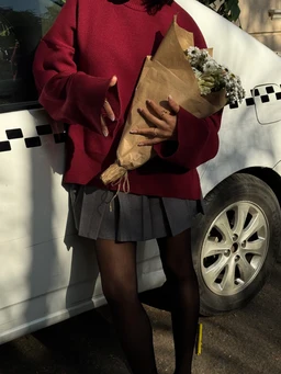 - What it is: A gray pleated mini skirt paired with a red oversized sweater. The model holds a bouquet of flowers, standing beside a white vehicle.
- Intended audience: Women (based on the skirt length, tights, and feminine styling).
- Notable attributes: Gray pleated fabric, short/mini length, dark sheer tights, and a chunky red knit sweater. Overall style is casual yet chic, with a slightly preppy, feminine vibe.