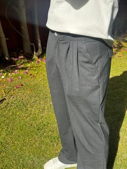 These are a pair of wide-leg trousers in a grey wool fabric, shown on a person from the waist down to the feet. The styling reads as menswear or unisex, with a relaxed yet tailored fit, featuring a flat front, pressed pleats, and a mid-rise waist. Visible attributes include the smooth wool texture, subtle sheen, front pockets, and a clean, straight leg silhouette.