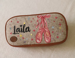 - Product: A rectangular pencil case with a ballet-themed design.
- Audience: Likely unisex or girls’ use, inferred from delicate ballet slippers and pink accents.
- Materials/attributes: Burlap-like fabric exterior with a brown trim and zipper; printed illustration of pink ballet pointe shoes with a bow, scattered colorful dots, and a small circular emblem. The visible text says “Laila.”