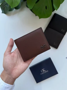 - What the product is: A compact brown leather wallet with a subtle textured grain, featuring a small metal CK logo on the front.
- Apparent audience: Unisex (suitable for men or women, based on the classic, minimalist bi-fold design and neutral brown color).
- Notable attributes visible: Brown textured leather, compact bi-fold style, small metallic CK emblem on the front; in the background there are dark and navy display boxes with CK branding.