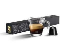 This image shows a single-serve espresso coffee capsule system with a small glass cup of dark espresso. Appears to target adults (likely men or unisex) given the sleek, minimal styling and compact cup. Notable attributes include a glossy black capsule, a clear glass cup with visible crema, and a dark, rectangular box packaging in black with gold branding.