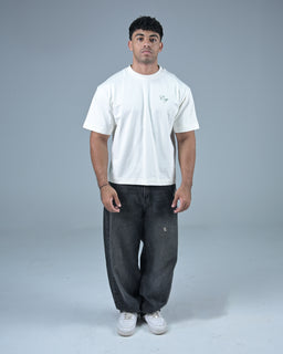 - A plain white short-sleeve T-shirt is shown on a male model, styled in a casual, relaxed fit with a standard crew neckline. The shirt features a small graphic or logo at the left chest.

- Audience: men (adult male model) judging from the cut, fit, and styling.

- Notable attributes: white cotton knit fabric, short sleeves, relaxed/tunic-like straight hem, paired with dark washed loose-fit jeans and white sneakers for a casual streetwear look. No visible branding beyond a small chest graphic.
