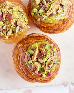 A photo of three pastry cruffins topped with chopped pistachios and dried fruits. The visible audience is unisex, judged from the pastries’ neutral, pastel-tinted, artisanal style. Notable visible attributes: flaky, golden-brown laminated pastry layers; green pistachio pieces, almonds or similar nuts, and small chunks of dried fruit on top, with a pistachio-green filling or glaze. Overall style is artisanal, pastry-forward, rustic. No branding visible.
