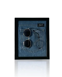 A framed display featuring a denim pocket from a blue jeans back pocket, with two pairs of circular sunglasses tucked into the pocket. The frame is black with a light reflection on the glass, and a small label tag is visible on the upper right of the pocket. The visible styling suggests unisex appeal. Notable materials: denim (jean fabric) and metal for the sunglasses; primary colors are blue denim and black frame.