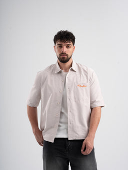 - What it is: A light brown/beige short-sleeve button-up shirt with subtle vertical stripes, shown worn over a white tee.
- Audience: Men's product (adult male model).
- Notable attributes: Lightweight woven fabric, soft neutral light brown color, short-sleeve shirt with a left chest pocket and a relaxed, casual fit, collared neckline.