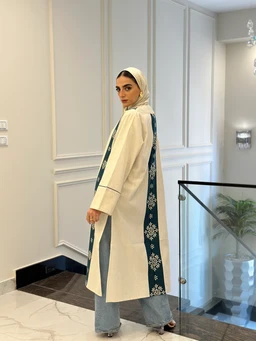 - What it is: A long, light-colored cardigan or overcoat with a dark vertical panel featuring a snowflake-like floral motif running down the sides.
- Intended audience: Women's (model wearing hijab suggests feminine styling).
- Notable attributes: Cream/ivory base fabric, wide cuffs, contrasting dark teal-blue side panels with white floral prints, open-front style, paired casually with light-wash jeans. Material appears knit or wool-like, with a tailored, architectural silhouette. Color palette is cream with dark teal panel accents. Style is modest, modern, and bohemian-inspired.